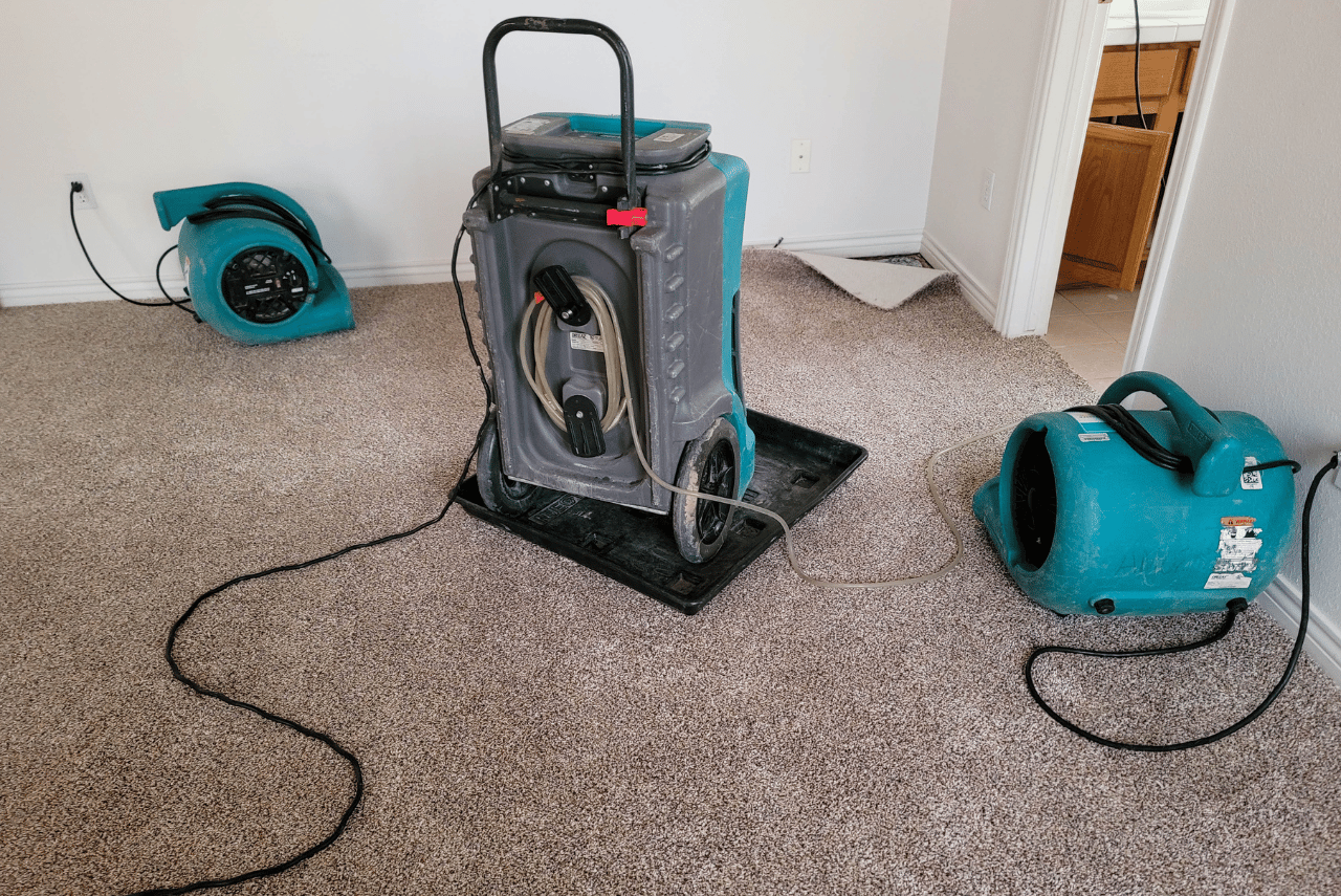 Three industrial air movers and a dehumidifier are set up on a carpeted floor in a mostly empty room, likely as part of water damage restoration or drying efforts.