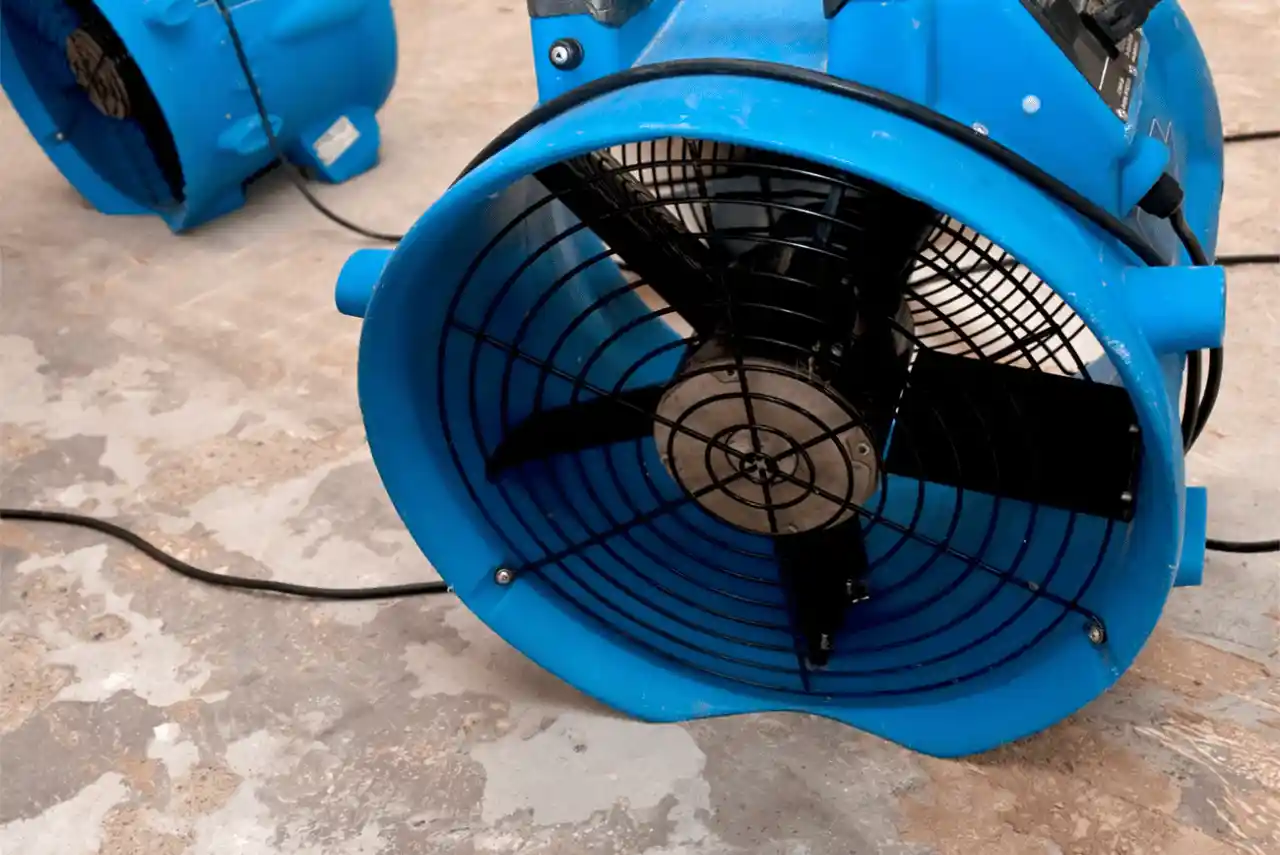 A large blue industrial floor fan sits on a concrete floor in Calgary, plugged in with a visible power cord—ideal equipment for a Water Damage Company. Another similar fan is partially seen in the background.