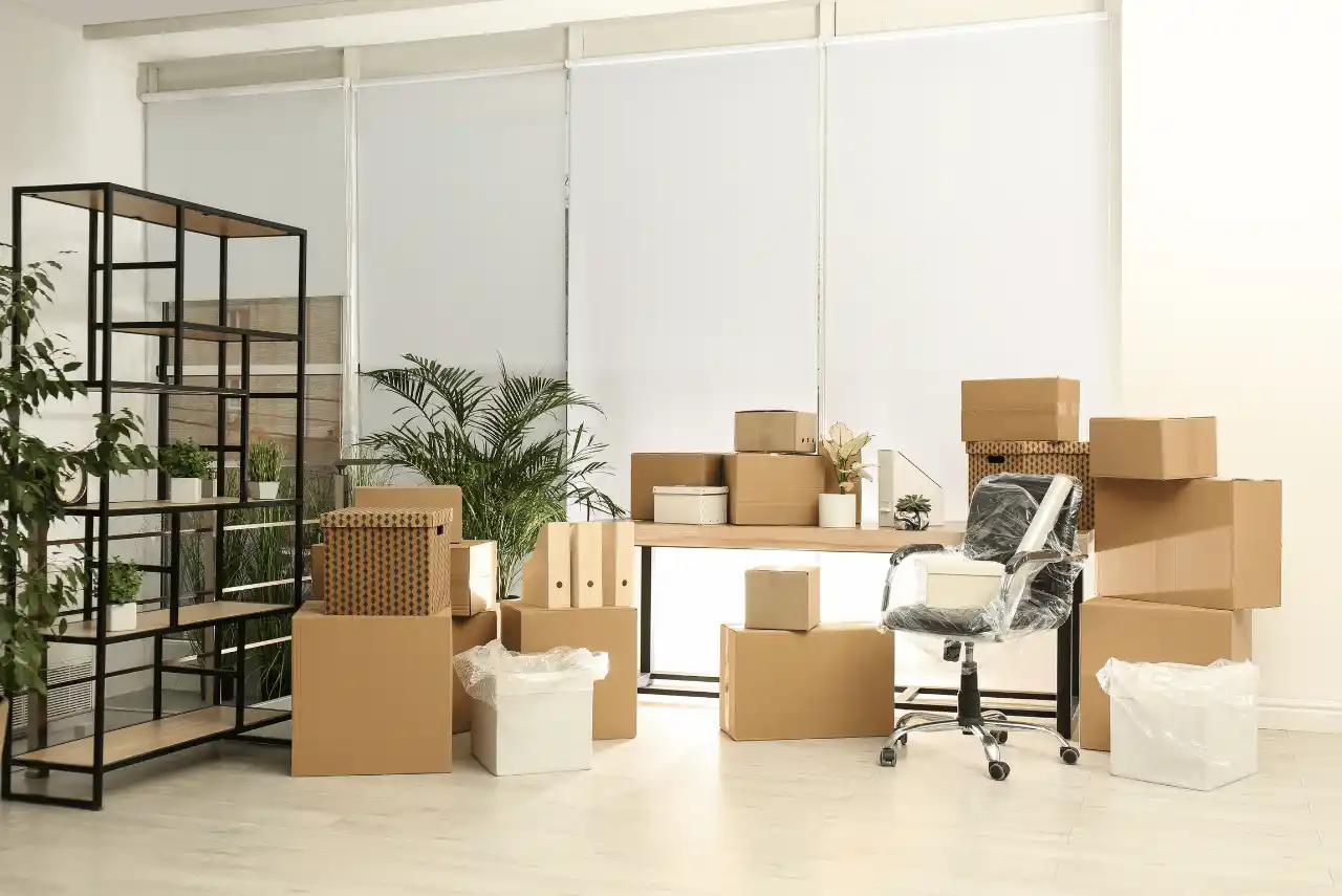 Office space with packed cardboard boxes, an empty bookshelf, a desk, and a wrapped office chair, hinting at relocation or content manipulation in progress.