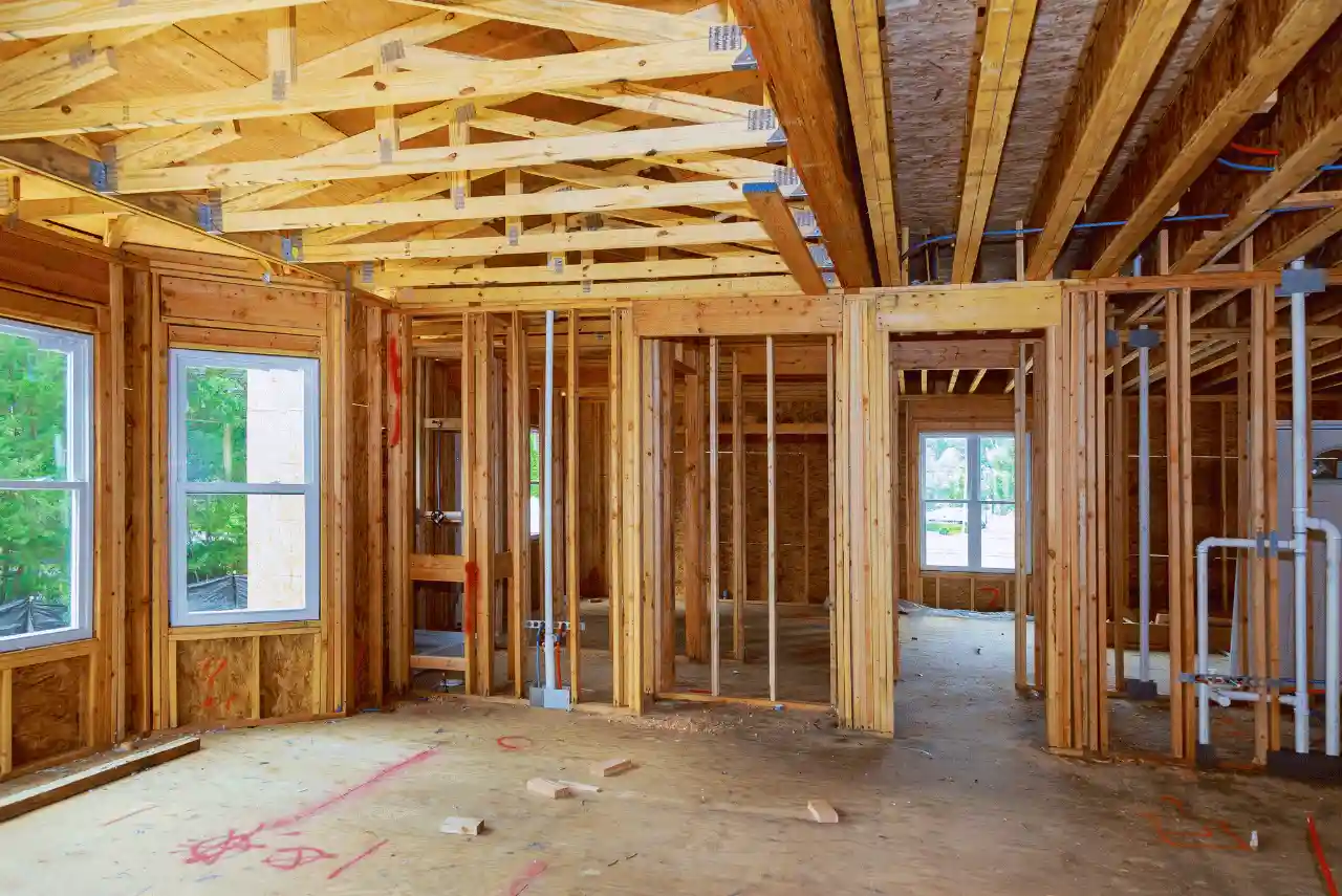 Interior view of a house under construction showing exposed wooden framing, unfinished walls, open windows, and visible ceiling beams with some plumbing and electrical work, highlighting ongoing structural repairs and detailed framing throughout the space.