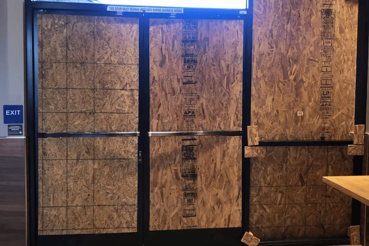 Double glass doors and nearby windows are covered with sturdy plywood, the work of professional board up services. A small sign reads "EXIT" on the left side. There is debris near the bottom right corner of the boards.