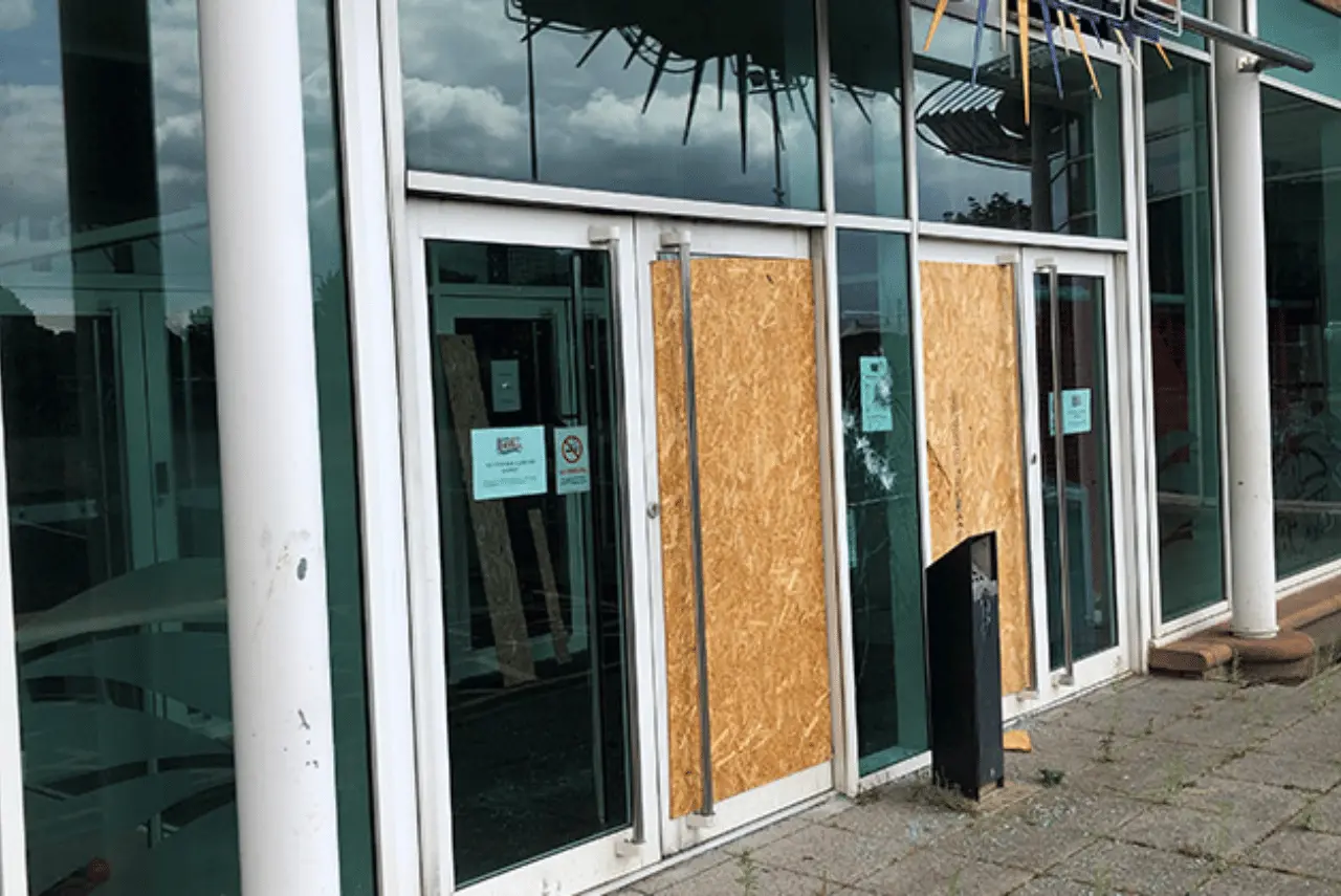 A glass storefront with two large windows and a door boarded up with plywood; signs posted on the glass; cloudy sky reflected in the glass.
