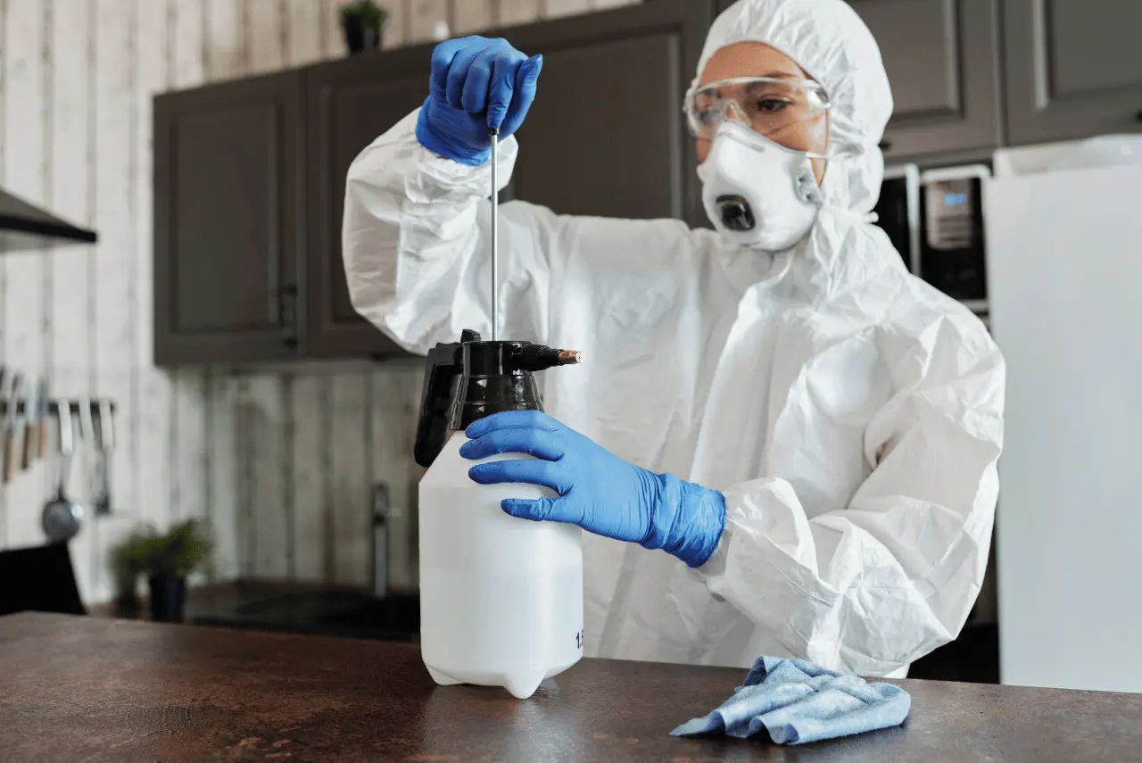 A person in protective gear and gloves prepares a disinfectant sprayer in a kitchen, with a cleaning cloth on the counter, exemplifying the high standards of Bio Cleaning in Calgary.