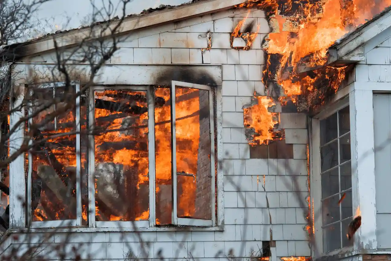 A house with white siding is on fire, with flames visible through open windows and burning the exterior wall—a situation where a skilled Calgary Restoration Company can make all the difference in returning your home to its original state.