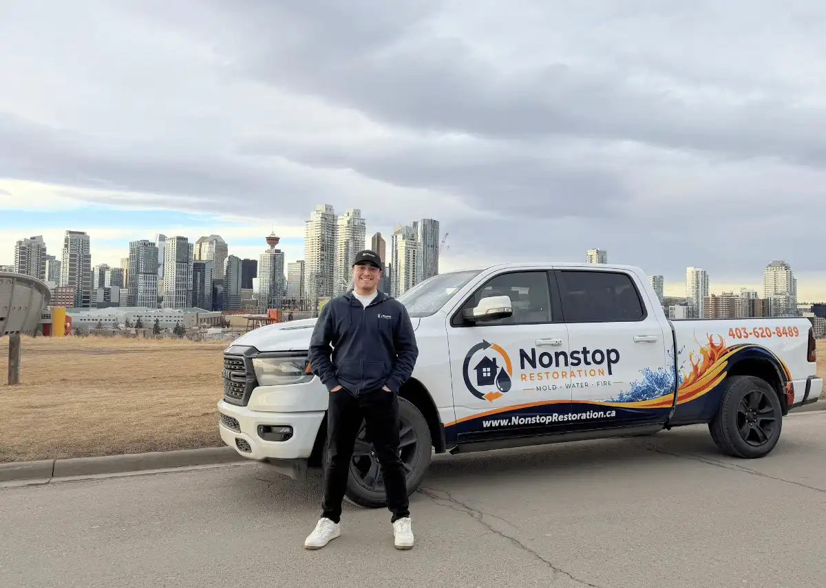 A person stands in front of a Nonstop Restoration truck on a roadside with a city skyline and cloudy sky in the background.