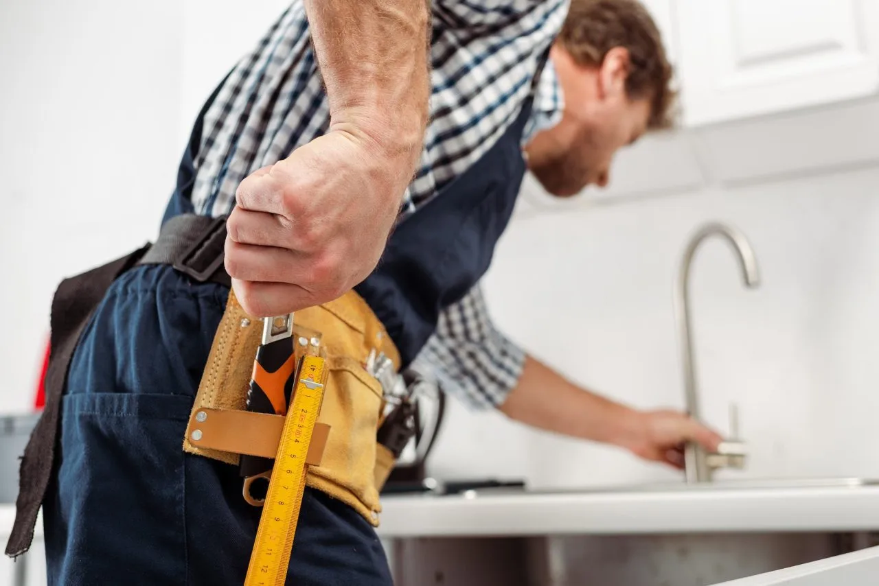 A man wearing a tool belt holds a ruler and checks a kitchen sink faucet, dressed in a plaid shirt and apron, with tools visible in the holster—ready for plumbing repairs or mold removal tasks.