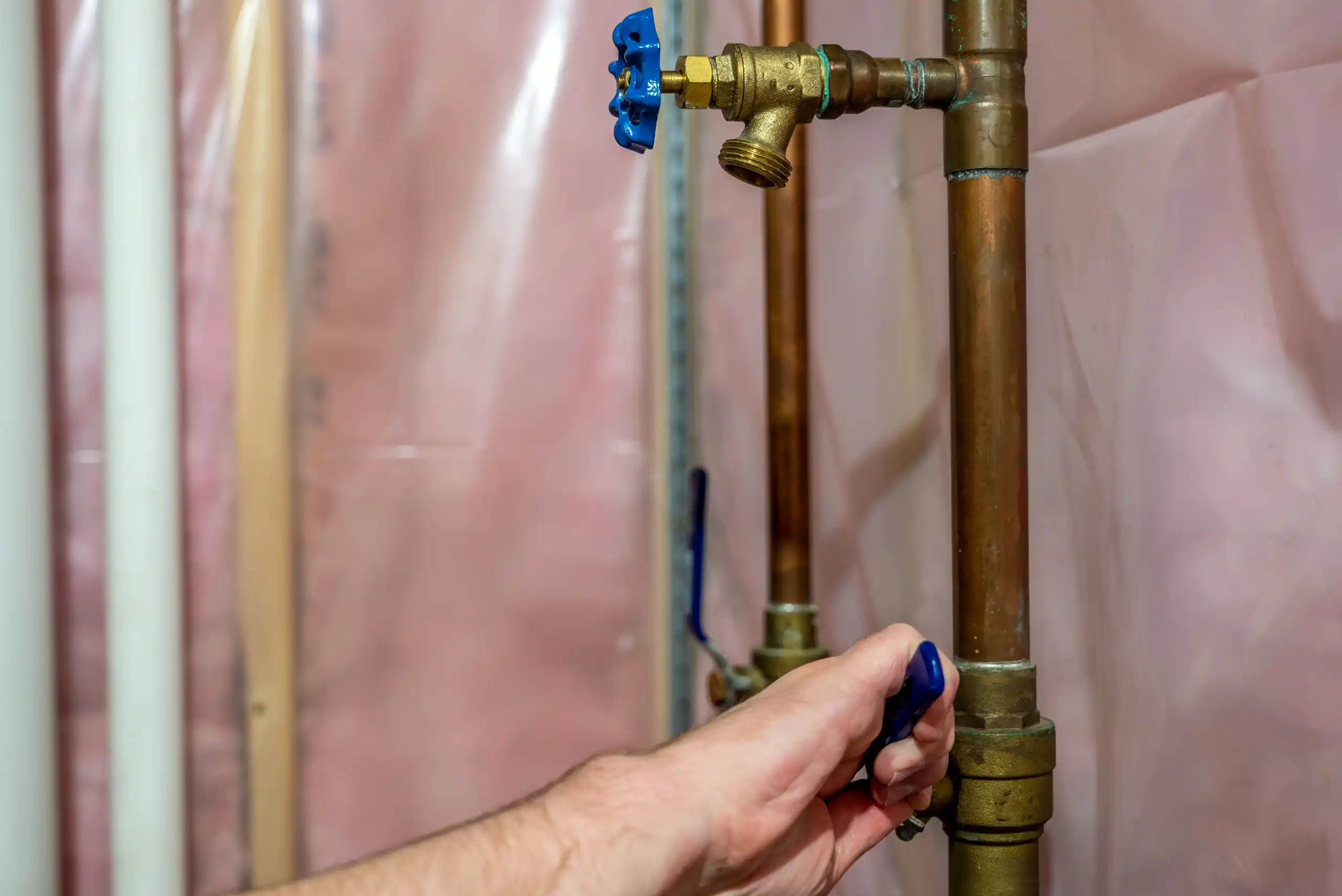 A hand turns a blue valve on a copper pipe mounted on a basement wall with pink insulation, demonstrating how to shut off water in case of an emergency leak.